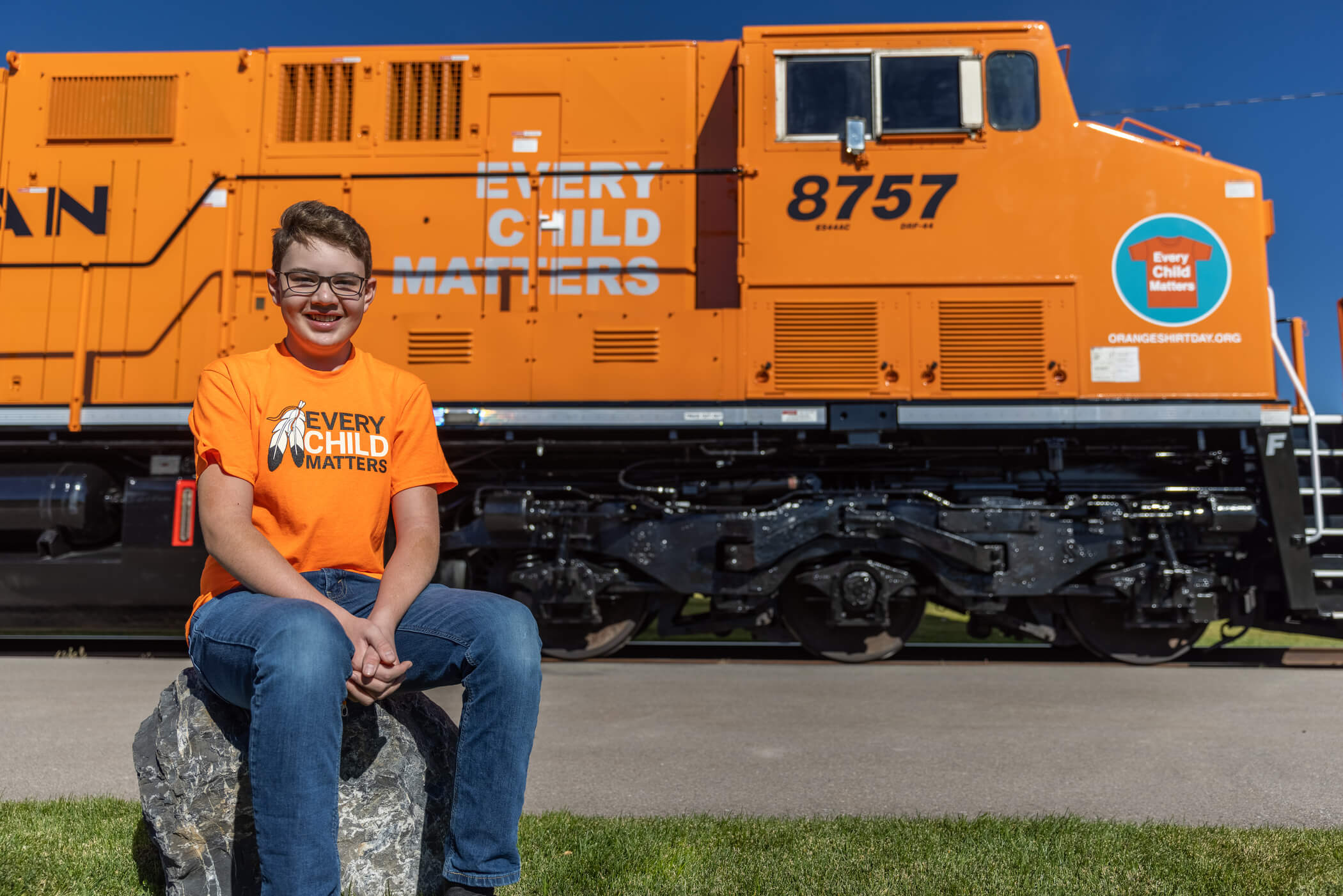A young person in an orange t-shirt sitting on a rock in front of a bright orange locomotive with "Every child matters" written on the side in grey letters.